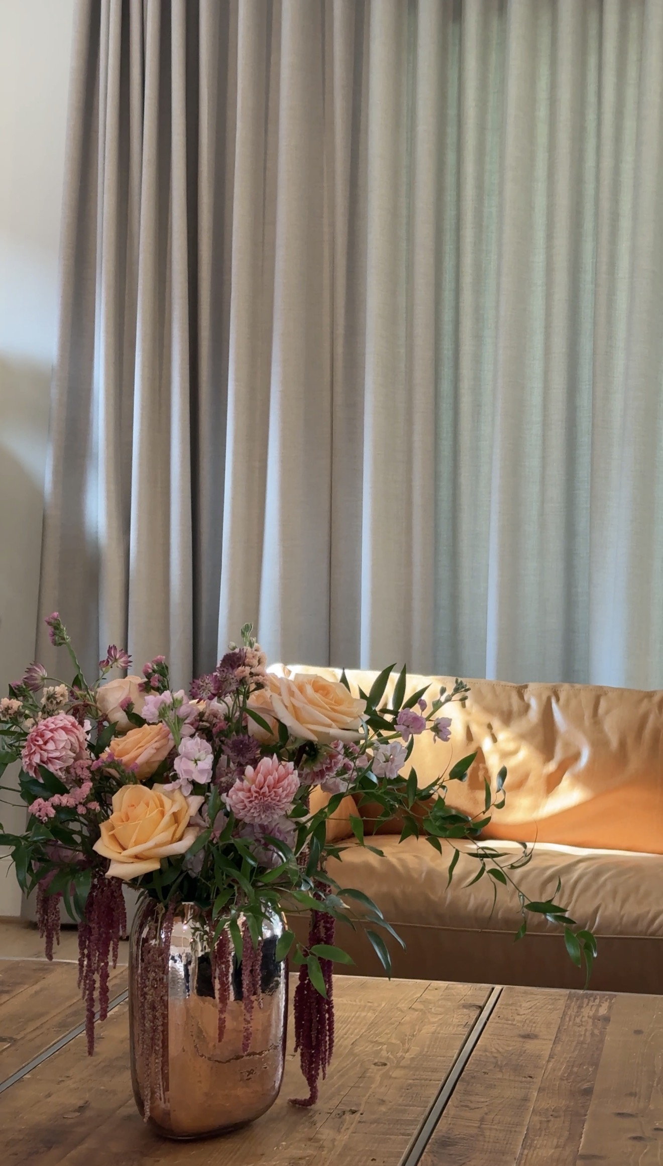 Peach roses and pink blooms with trailing burgundy amaranth, arranged in a copper-toned vase beside a warm living-room sofa