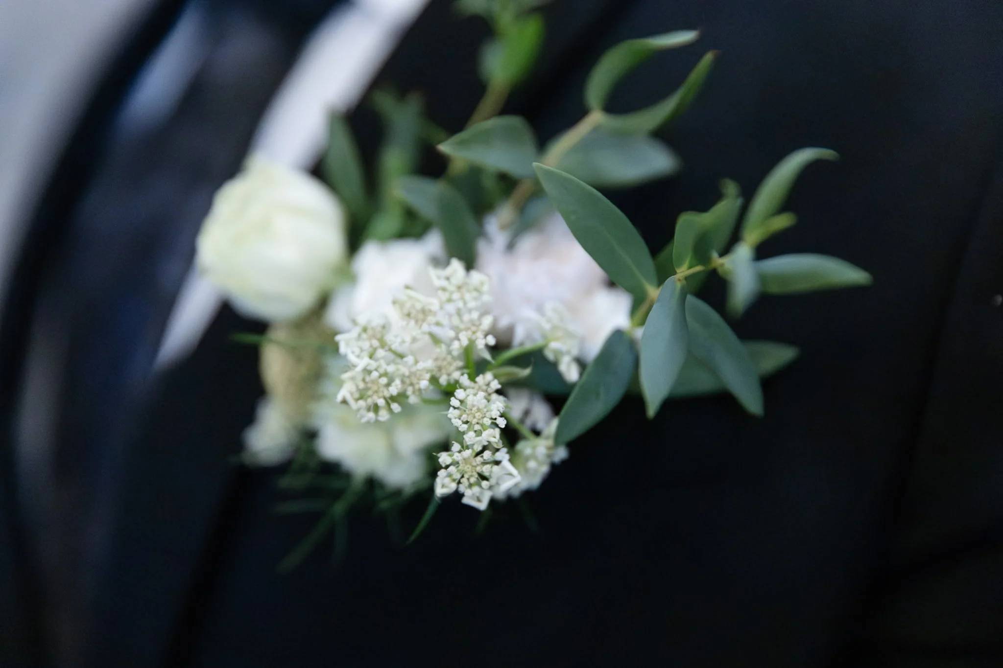 A white floral boutonniere with green foliage on a dark suit